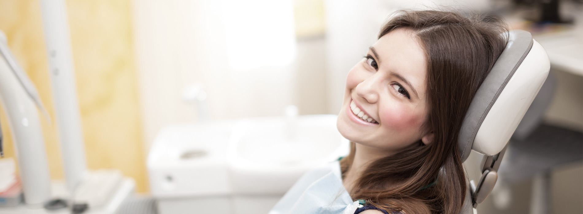 A person is seated in a dental chair, receiving care from a dental professional who stands behind them.