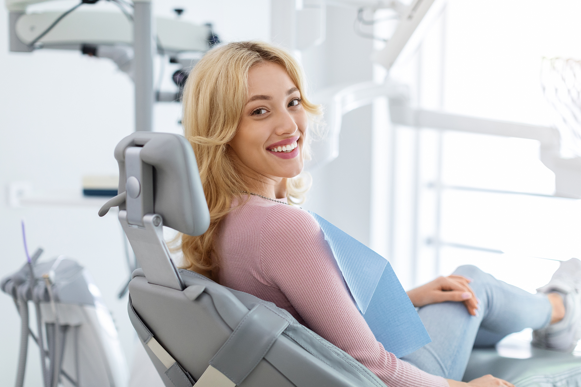 The image shows a woman sitting in a dental chair with her mouth open, wearing a blue apron, smiling at the camera, while an orthodontist s office setting is visible in the background.