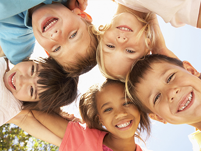 A group of children, both boys and girls, smiling together in a sunlit outdoor setting.